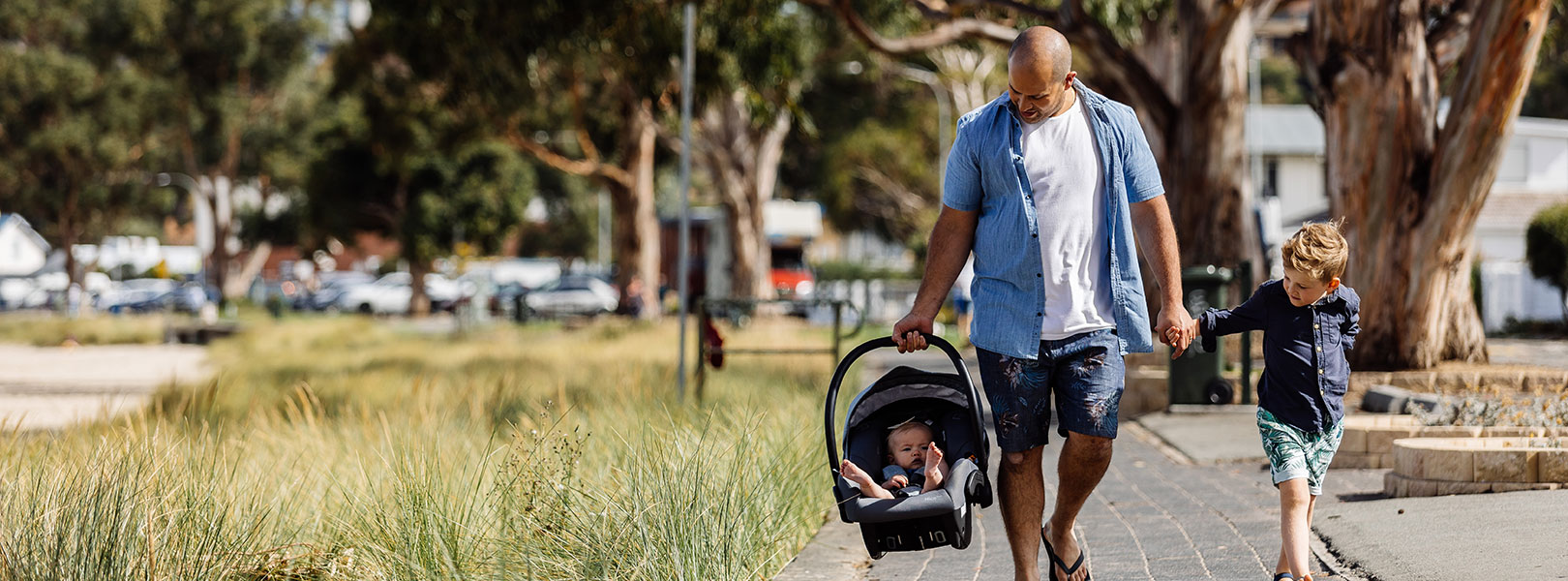 Dad walking along footpath holding toddlers hand and carrying baby in capsule.