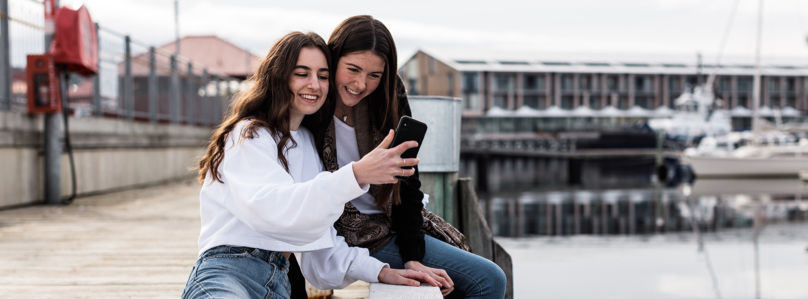 Teenage girls taking selfies on phone while sitting on dock.