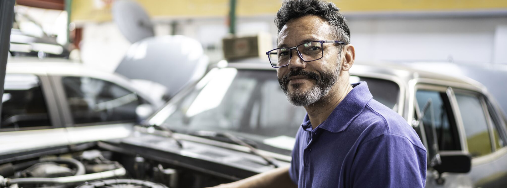 Male mechanic wearing blue shirt looking at camera with car bonnet open in background