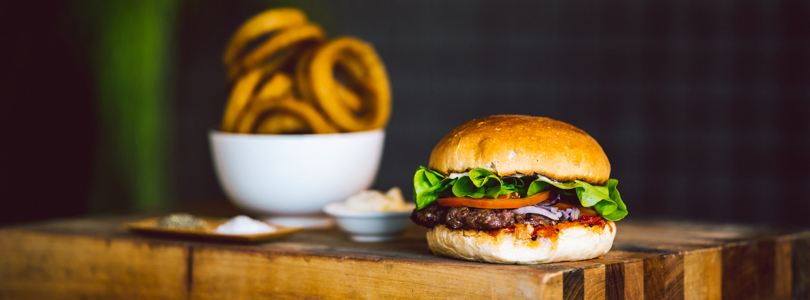 Image of a delicious beef burger sitting on a wooden counter next to an overflowing bowl of onion rings.