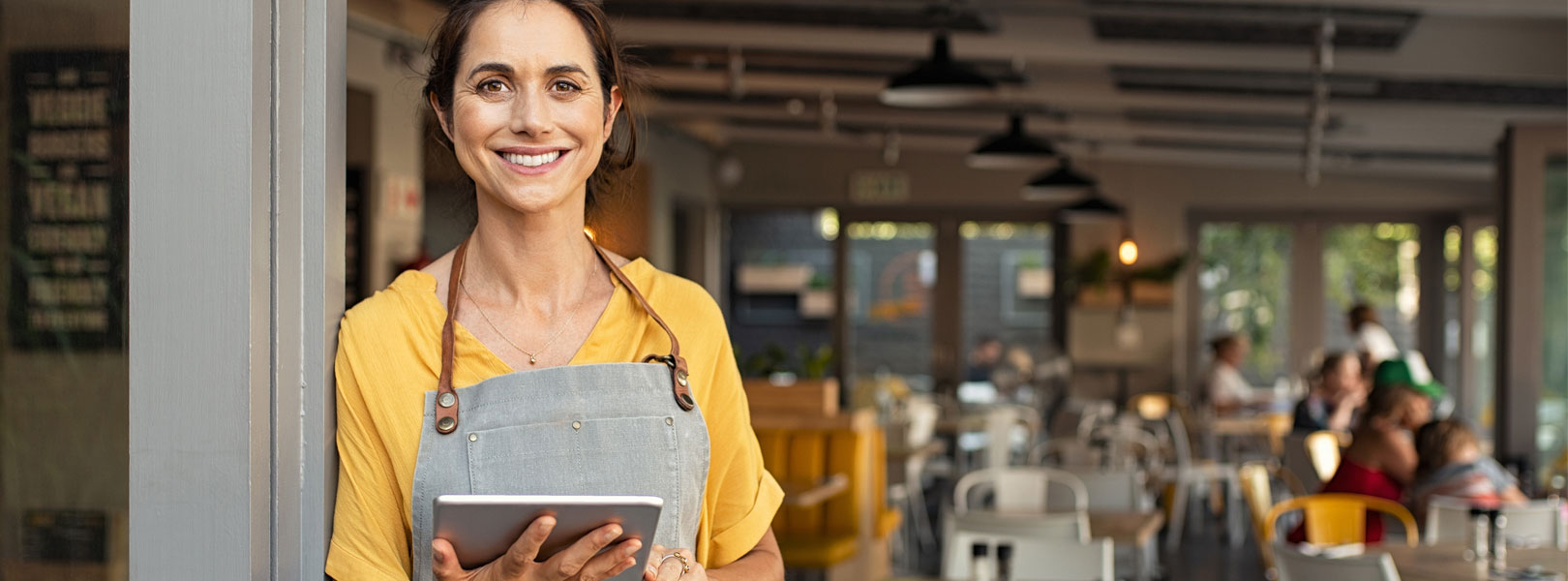 Female standing at front of cafe wearing a yellow shirt, grey apron and holding iPad with patrons blurred in background