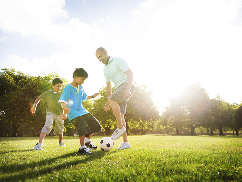 Dad playing soccer with children in the park on a sunny day.